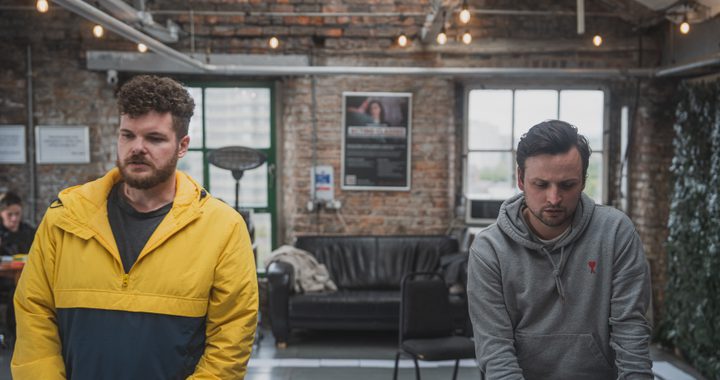 Two men looking sad. Man 1 has short curly hair and a beard he is wearing a yellow and man 2 has straight short hair and is wearingblue jacket and the other is wearing a grey jumper