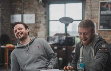 Two men sat at a table laughing, both wearing grey jumpers.