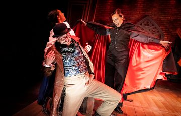 Image of Dracula in a red cape going towards 2 actors looking scared