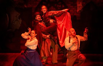 Image of 4 actors on stage with a red light, one of the actors is dressed as Dracula