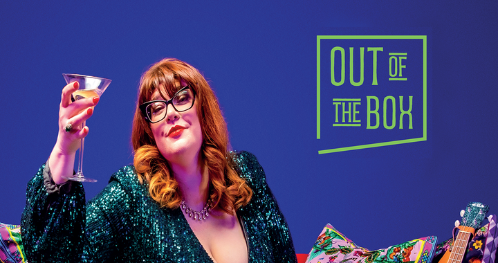 A woman with ginger hair, wearing glasses and a green sequin dress sat on a sofa with a martinin glass held up in the air