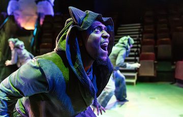 A man in a hood standing sideways and looking up with his mouth open. Two people standing backwards in the background. With a green light behind him and a blue light in front of him.