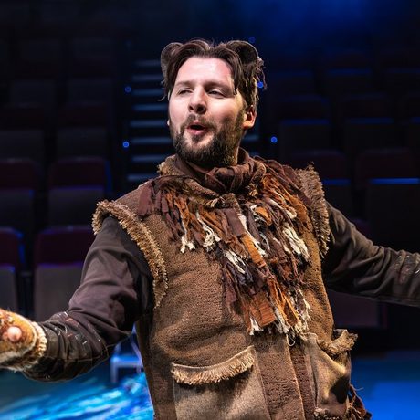 A man in a brown costume with his arms to the sides and blue lights in the background.