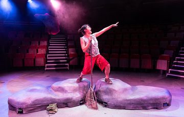 A woman with her left hand to the side holding a butterfly net upside down standing on two rocks. With pink and blue lights in the background.