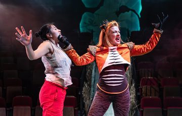 A woman standing sideways with her arms spread. Another woman holding her by the chin. With red lights in the background.