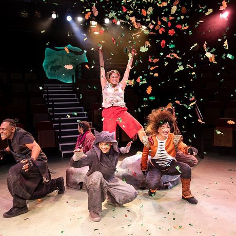 A woman standing on a rock with her hands up. Five people squatting around the rock with orange and green confetti flying around them and yellow, white, and red lights in the background.