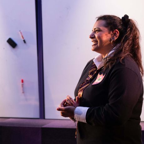 A brown women, with curly hair, tied into a pony tail. Wearing white shirt and a school jumper.
