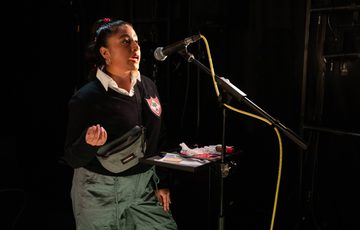 A young brown woman hair tied back with a braid, wearing a black top and green trousers and a crossbody bag talking into a mic.