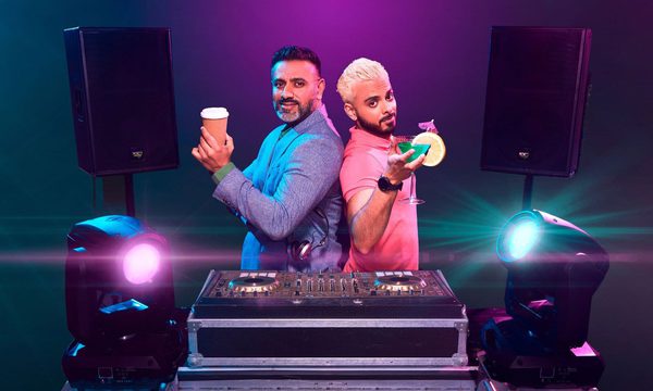 2 Asian men stood behind a DJ set. The older man has a coffee and the younger man has a cocktail.