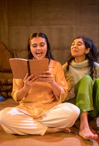 Two girls sat on the floor, one absorbed in a book while the other is listening attentively.