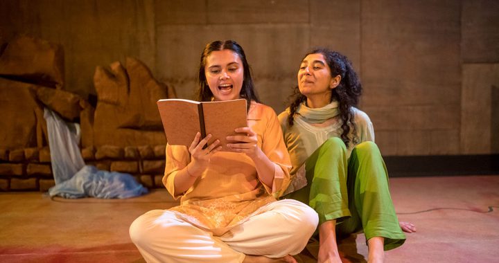Two girls sat on the floor, one absorbed in a book while the other is listening attentively.