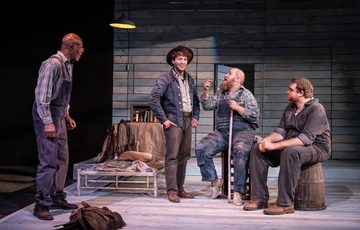 Four actors are gathered onstage discussing an important topic. The two actors on the left are stood, the two actors on the right are sat as the conversation takes place.