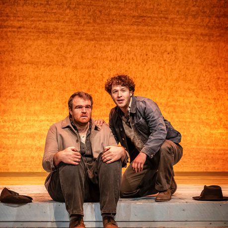William Young who plays Lenny is sat on the floor holding his knees whilst Liam King who plays George kneels beside him.