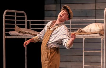 The actor is shown onstage, in a shirt, dungarees and flat cap. He looks as though he is in the middle of shouting something to another person.