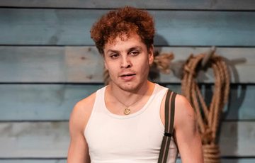 The actor is shown standing onstage wearing a vest, on glove, and one brace up over his shoulder