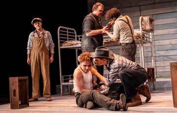 Five actors are shown onstage. The two upstage are crouched and appear to be having a serious discussion. Two actors are stood behind them, one has a cut hand and the other holds a rag attempting to stop the bleeding. The final actor is stood to the left of the screen and is onlooking.