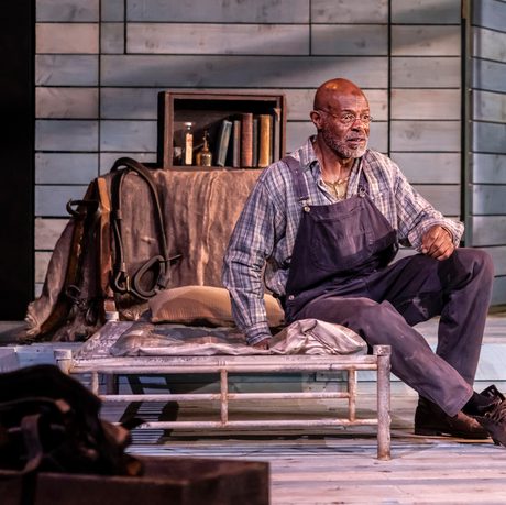 Jeff Alexander, who plays Crooks is sat on a step wearing a long sleeve shift and dungarees.