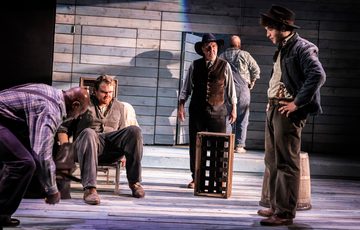 The actors are spread around the stage. The focus is one actor sat in a chair. He looks angry, confused and serious