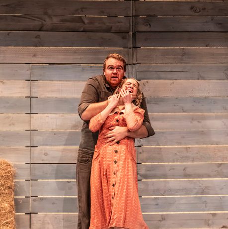 Two actors are shown on stage. One actor has a hold of the other from behind and appears to be restraining her out of fear.