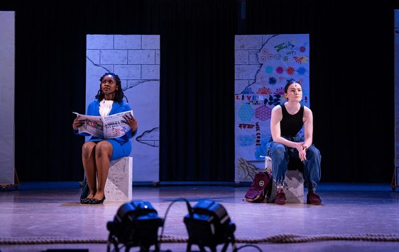 Two actors on stage sat on blocks. They look ahead into the distance