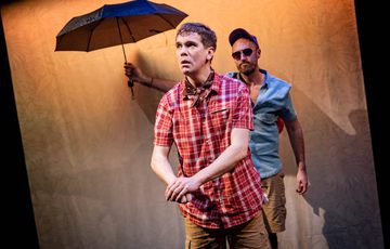 The actors are on stage. The actor downstage looks worried, whilst the actor upstage is wearing a cap, sunglasses and holding an opened umbrella.