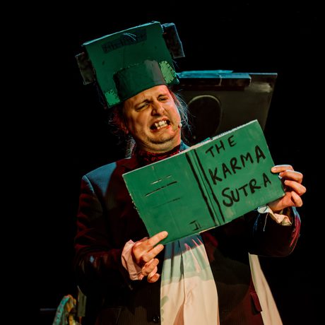 Actor is shown wearing a Frankenstein inspired head piece holding a book titles 'The Karma Sutra'