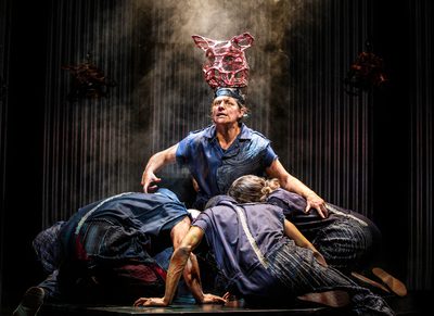 A person with a headpeice representing a pig kneeling on floor with a group of people hurdled around with their heads down