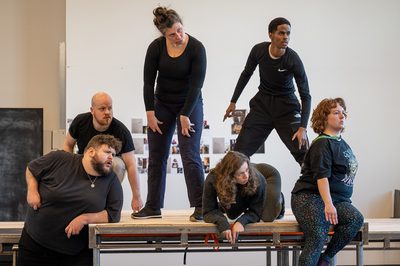 6 men and women on a stage in a variety of positions, looking out to the distance with intrigued expressions.