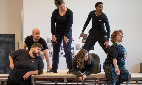 6 men and women on a stage in a variety of positions, looking out to the distance with intrigued expressions.