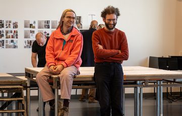 One man sat on a table smiling and a man stood next to him with his arms crossed and laughing.