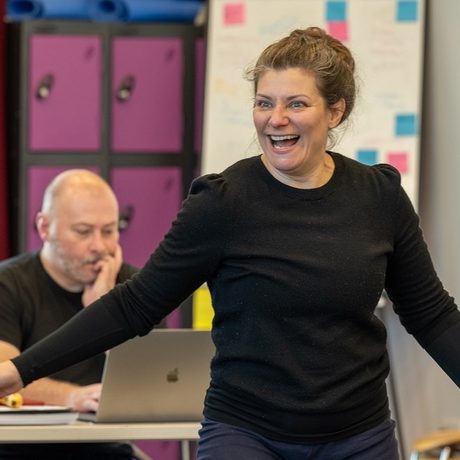 A woman stood with her arms out smiling. A man sat at a desk behind her looking at a laptop.