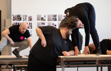 a man and woman on a stage on different levels, leaning in to each other. A man in the background of the stage squatting down watching them and a woman lay on stage half out of shot looking up to the ceiling.