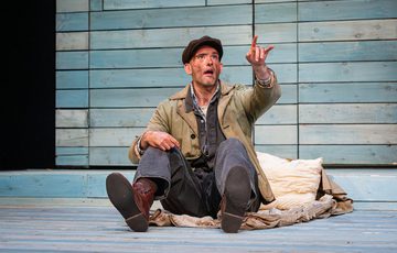 An actor sits on the stage and points up to the sky in the distance