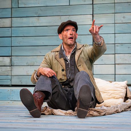 An actor sits on the stage and points up to the sky in the distance