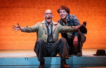 Two actors are on stage. One is sat on a step, the second is crouched behind him. The actor sat down appears to be explaining something, the actor behind him looks ahead and smiles.