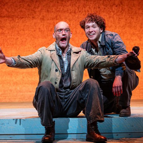Two actors are on stage. One is sat on a step, the second is crouched behind him. The actor sat down appears to be explaining something, the actor behind him looks ahead and smiles.