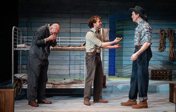 Luke Richards, playing Lennie stands with his head in his hands behind Liam King playing George as he talks to Laurence Pears playing Slim