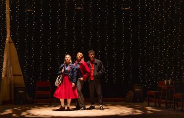 3 actors stand centre stage under a spotlight looking ahead into the distance.