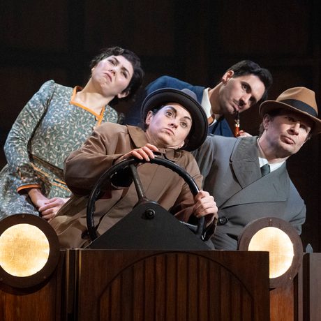 Four actors are shown to be in a car on stage. They are all leaning to stage left as if the car is turning.