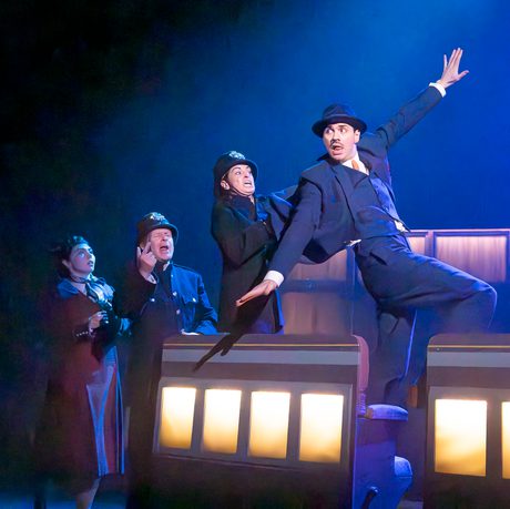 Four actors are shown onstage. The actor towards the front is doing a dramatic pose whilst the others watch him