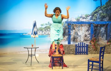 The actress is in front of a Greece setting, she stands on top of a chair and is about to jump off of it.