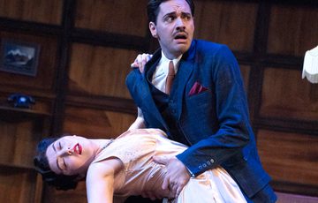 Two actors are on stage. One looks as though they have fainted whilst the other holds themup.