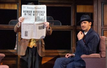 Two actors are on stage. On of them is stood up with a newspaper in front of their face. The other is sat in an armchair looking out into the distance.