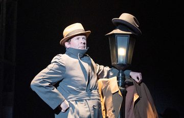An actor is shown in a top hat and long coat leaning on a Victorian style lamp post