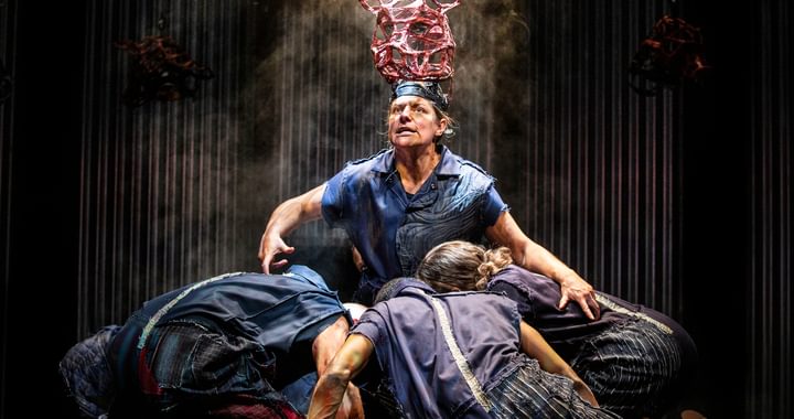 A person with a headpeice representing a pig kneeling on floor with a group of people hurdled around with their heads down