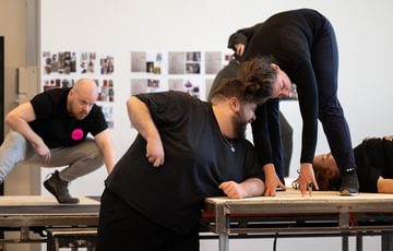 a man and woman on a stage on different levels, leaning in to each other. A man in the background of the stage squatting down watching them and a woman lay on stage half out of shot looking up to the ceiling.