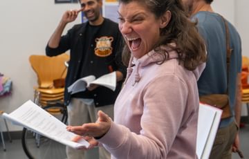 A woman stood looking to her side and pointing, with a big smiling gesture on her face. Two men stood behind her, one laughing.