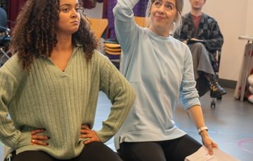 Two women sat on a luggage trunk. One with hands on hips another holding a script in one hand and other hand on her head.