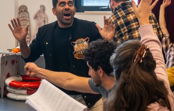 Man stood up with his hands in the air looking scared with two other people sat with back to camera waving.