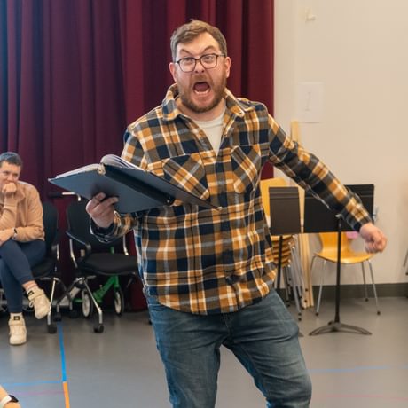 A man stood in a rehearsal room with a script in his hand pulling a scared expression.